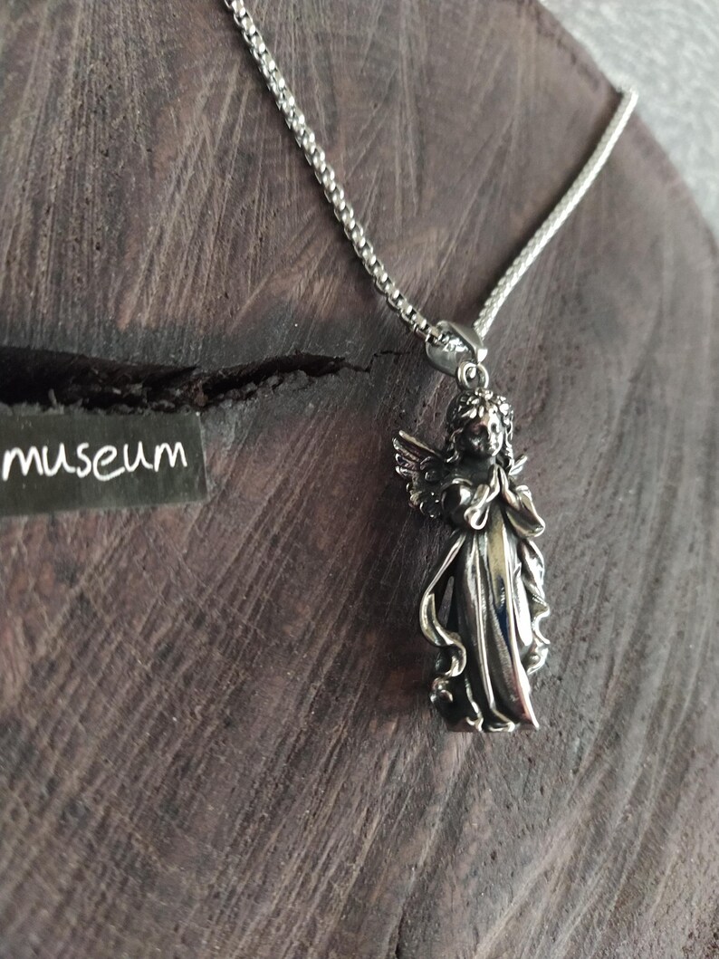 May include: A silver chain necklace with a pendant of a silver angel with wings outstretched. The angel is wearing a flowing robe and has a serene expression on its face.