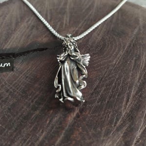 May include: A silver necklace with a detailed angel pendant. The angel is depicted with wings and a flowing gown.