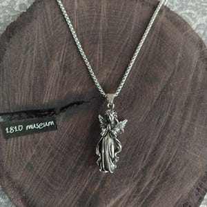 May include: A silver chain necklace with a pendant of a praying angel. The angel is detailed with wings and a flowing robe. The necklace is on a wooden surface with a tag that reads "1810 museum".