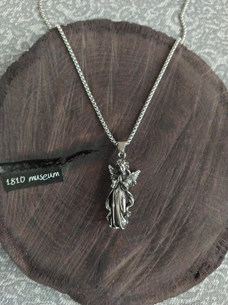 May include: A silver chain necklace with a pendant of a praying angel. The angel is detailed with wings and a flowing robe. The necklace is on a wooden surface with a tag that reads "1810 museum".