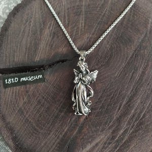 May include: A silver chain necklace with a pendant of a silver angel with wings. The angel is holding its hands together in prayer. The necklace is on a dark brown wooden surface.