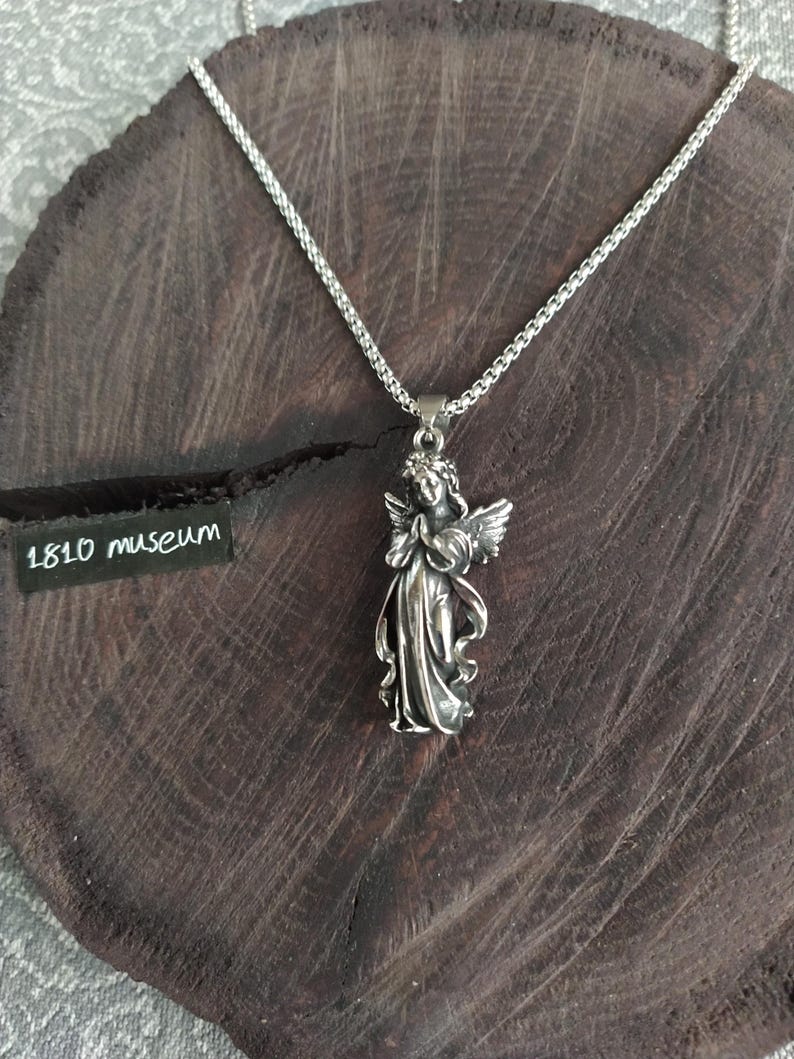 May include: A silver chain necklace with a pendant of a silver angel with wings. The angel is holding its hands together in prayer. The necklace is on a dark brown wooden surface.