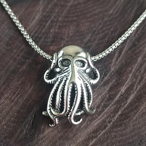 Könnte beinhalten: Halskette mit einem silberfarbenen Anhänger in Form eines Oktopus. Der Anhänger hat detaillierte Tentakel und einen stilisierten Kopf und hängt an einer feinen Kette. Die Kette wird vor einem dunklen Holzmaserungshintergrund gezeigt.