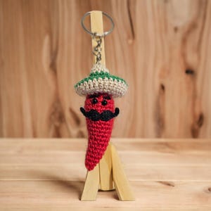 May include: A red crocheted chili pepper with a green and white sombrero, black moustache, and googly eyes. The chili pepper is attached to a key chain.