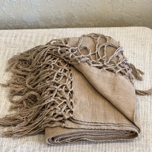 Puede incluir: Una manta de punto marrón con detalles de flecos. La manta está doblada y tiene una textura tejida.