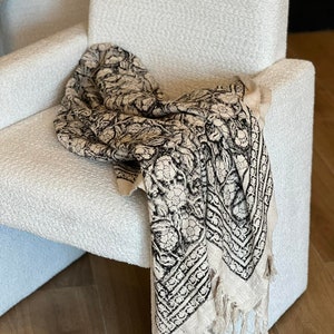 Könnte beinhalten: Ein weißer Sessel mit einer schwarz-weißen Musterdecke darüber gelegt. Die Decke hat einen Fransenrand und ein florales Design.