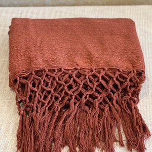 Op de afbeelding: Een geweven wollen deken in roestkleur met een franje rand. De franje is gemaakt van een geknoopt macramé-patroon.