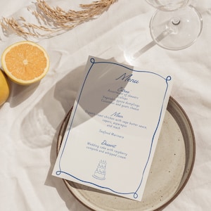 Może przedstawiać: Biała karta menu z niebieskim tekstem i niebieską ramką. Menu zawiera opcje przystawek, dań głównych i deserów. Przystawka obejmuje przegrzebki, pierożki gyoza oraz ogórek i kozi ser. Danie główne to ekologiczny kurczak pieczony z sosem maślanym z szałwią, kaparami, szparagami i puree. Deser to tort weselny z kompotem malinowym i bitą śmietaną.