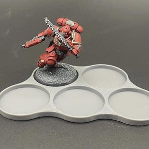 May include: A red and gray miniature figure of a space marine with a chainsword, posed on a gray, multi-holed tray. The tray is designed to hold multiple miniatures, with a textured base for the figure.