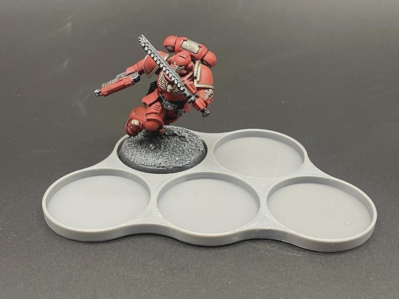 May include: A red and gray miniature figure of a space marine with a chainsword, posed on a gray, multi-holed tray. The tray is designed to hold multiple miniatures, with a textured base for the figure.