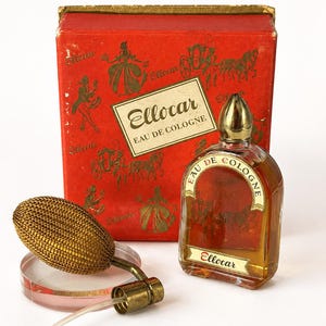Puede incluir: Juego vintage "Ellocar Eau de Cologne". El juego incluye una caja roja con ilustraciones doradas, un frasco de perfume de vidrio con tapa dorada y un atomizador dorado. La botella está llena de un líquido ámbar claro. La caja y la botella están etiquetadas "Ellocar".