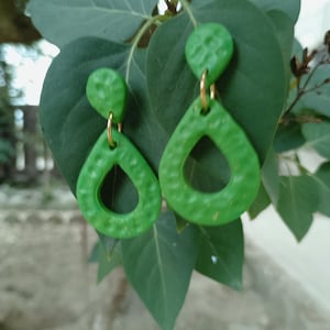 Peut inclure: Une paire de boucles d'oreilles en forme de goutte d'eau vert vif, texturées, avec des cerceaux dorés. Les boucles d'oreilles sont suspendues à une feuille verte, mettant en valeur leur couleur et leur design. Elles ont une texture martelée.