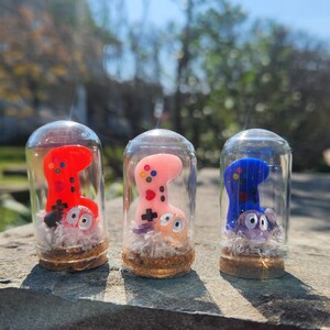 May include: Three glass jars with cork tops, each containing a small, colorful, hand-painted figurine of a video game controller. The figurines are red, pink, and blue.