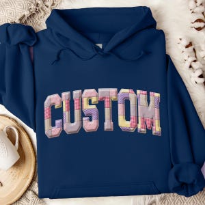 Puede incluir: Sudadera con capucha azul marino con la palabra "CUSTOM" en el pecho. Las letras están diseñadas con un patrón de patchwork en tonos rosa, morado, amarillo y gris. La sudadera tiene una capucha con cordón y un bolsillo delantero.