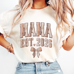 Può includere: Felpa color crema con la scritta "MAMA EST. 2026" in un carattere marrone, simile al plaid. Sotto il testo c'è una grafica a fiocco marrone. La felpa è in stile casual.