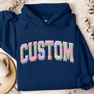 Puede incluir: Una sudadera con capucha azul marino con la palabra "CUSTOM" en un diseño de patchwork en el pecho. Las letras están rellenas de cuadrados y rectángulos de colores pastel, dando una apariencia acolchada. La sudadera tiene una capucha con cordón y un bolsillo delantero.