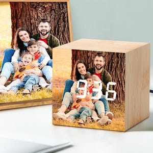 May include: A wooden cube clock showing the time 9:36 with a family photo printed on its sides. The clock has a light wood grain finish and a digital display. A framed family photo is visible in the background.