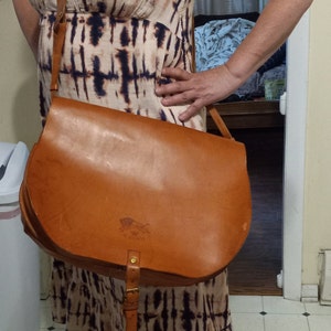 May include: A woman wearing a white and brown tie-dye dress with a brown leather satchel bag with a brass buckle. The bag has a small embossed logo on the front.