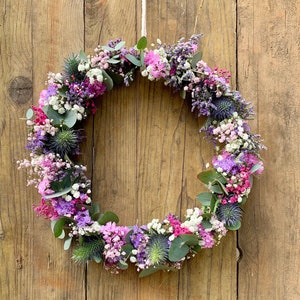 Könnte beinhalten: Ein Blumenkranz aus rosa, lila und weißen Blüten, Grün und Eukalyptuszweigen. Der Kranz hängt an einer Holztür.