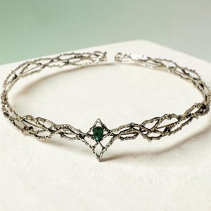 May include: A silver-toned tiara with a delicate, intertwined branch design. The tiara features a central, emerald-green gemstone set within a diamond-shaped frame. The open design is elegant and suitable for special occasions.