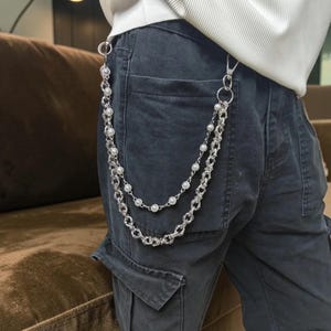 May include: A silver-colored chain with faux pearls and a decorative chain design. The chain is attached to a pair of dark gray cargo pants. The chain is hanging down from the pants.