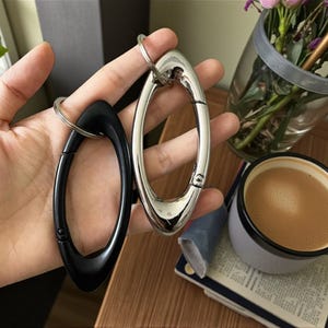 Könnte beinhalten: Zwei ovale Karabiner-Schlüsselanhänger, einer schwarz und einer silberfarben, werden in einer Hand gehalten. Die Schlüsselanhänger haben ein federbelastetes Tor für eine einfache Befestigung. Eine Tasse Kaffee und eine Vase mit Blumen sind im Hintergrund.