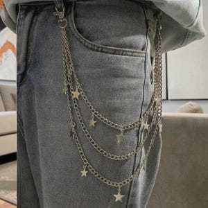 Op de afbeelding: Zilverkleurig multi-keten riemaccessoire met sterbedels bevestigd aan een lichtblauwe denim jeans. De kettingen hangen in drie lagen, met stervormige bedels die aan elke ketting bungelen.
