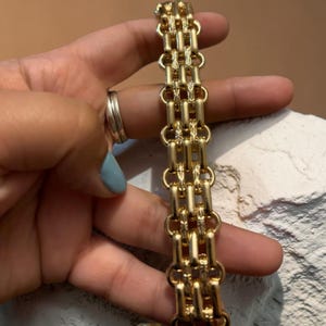 Könnte beinhalten: Ein goldfarbenes Armband mit einem Gliederdesign. Das Armband besteht aus mehreren Reihen rechteckiger Glieder, die durch kleinere, dekorative Glieder verbunden sind. Das Armband wird vor einem neutralen Hintergrund gehalten.