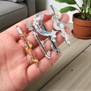 Könnte beinhalten: Eine Sammlung detaillierter Metallschwert-Anstecknadeln. Eine Nadel zeigt ein rotes und gelbes Flammendesign, während andere blaue, schwarze und silberne Akzente aufweisen. Die Anstecknadeln werden vor einem neutralen Hintergrund präsentiert, um ihre komplizierten Designs hervorzuheben.