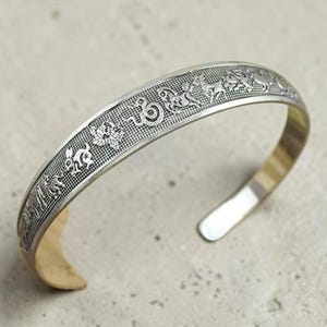 Könnte beinhalten: Silberfarbenes Armreif-Armband mit detaillierten Tier- und Symbolgravuren. Das Armband hat ein strukturiertes Band mit goldfarbenem Innenraum. Das offene Design ermöglicht ein einfaches Tragen. Ein einzigartiges Schmuckstück.