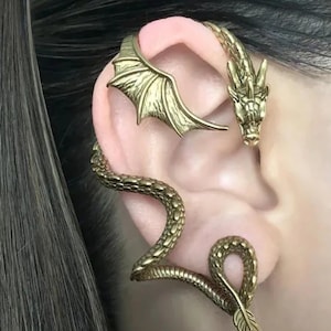 Könnte beinhalten: Ein goldfarbener Ohrschmuck mit Drachen- und Schlangenmotiv. Der Ohrschmuck umschließt das Ohr mit einem Drachenkopf, einem Flügel und einer sich windenden Schlange. Der Schmuck ist detailliert und verziert.