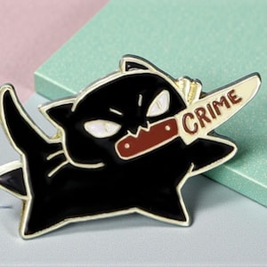 Könnte beinhalten: Schwarzer Katzen-Emaille-Pin mit goldenem Umriss, der ein Messer mit dem Wort "CRIME" hält. Die Katze hat weiße Augen und einen bedrohlichen Ausdruck. Der Pin befindet sich auf einem hellblauen und rosa Hintergrund.