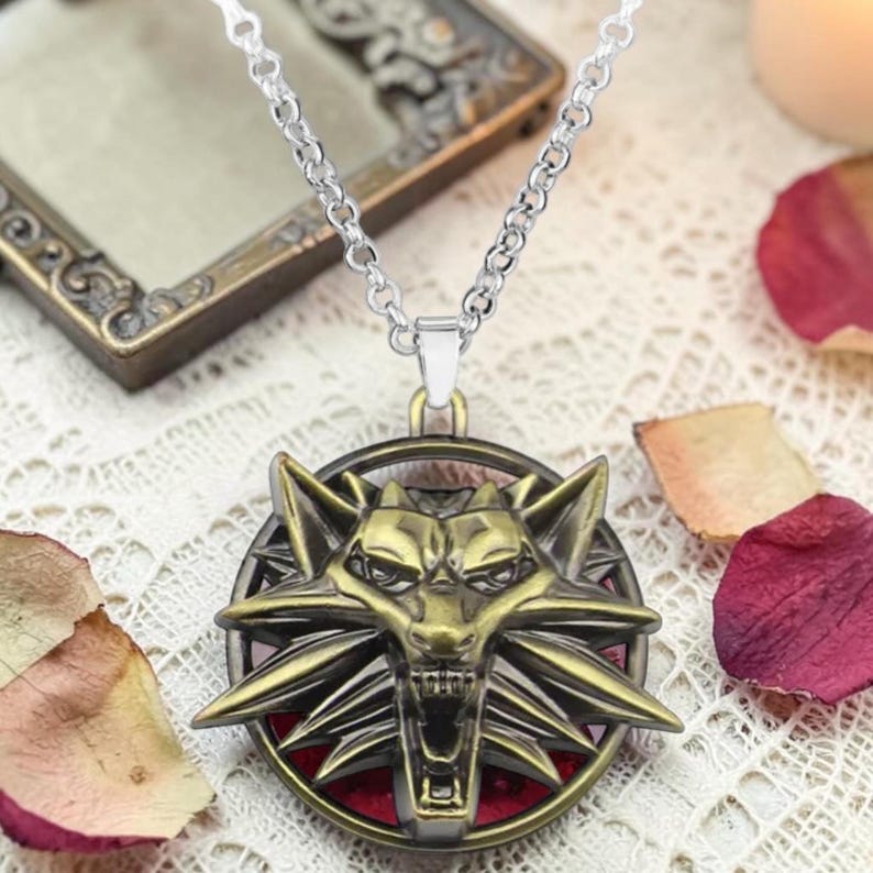 Puede incluir: Collar de cadena plateada con un colgante con un emblema de cabeza de lobo de color bronce. El colgante tiene un fondo rojo y est&aacute; rodeado por un marco circular. El collar se muestra sobre una superficie de encaje blanco.