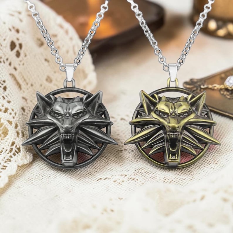 Puede incluir: Dos collares con colgante de metal. Uno es de color gris plomo, el otro dorado. Cada uno presenta un emblema de cabeza de lobo dentro de un marco circular, suspendido de una cadena plateada. Los colgantes est&aacute;n detallados con &aacute;ngulos agudos y una expresi&oacute;n feroz.