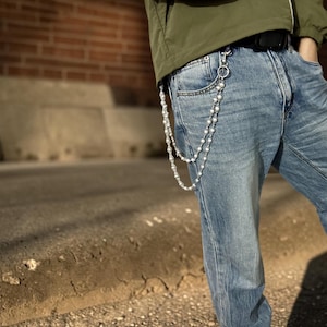 May include: Light-wash denim jeans with a pearl chain attached to the belt loop. The chain is made of small, round, white pearls. The person is wearing a green jacket and a black belt. The background is a brick wall and a concrete sidewalk.