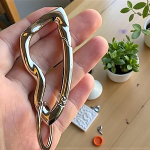 Könnte beinhalten: Ein glänzender, silberfarbener Karabinerhaken mit einem einzigartigen, wellenförmigen Design. Der Haken hat einen federbelasteten Verschluss und einen kleinen, runden Ring am unteren Ende. Dieser Artikel wird wahrscheinlich zum Befestigen von Schlüsseln oder anderen kleinen Gegenständen verwendet.