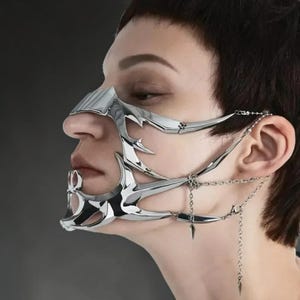 May include: A silver-colored face mask with a unique, spiky design. The mask covers the nose and mouth, with chain details connecting to the ear. The mask has a futuristic, avant-garde aesthetic.