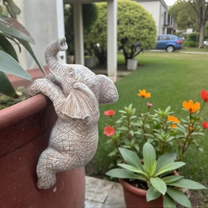 Könnte beinhalten: Eine detaillierte, strukturierte, graue Elefantenfigur, die am Rand eines terrakottafarbenen Blumentopfs hochklettert. Der Elefant hat seinen Rüssel erhoben und ist so positioniert, als würde er sich hochziehen. Im Hintergrund gibt es bunte Blumen und grünes Laub.