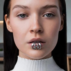 Könnte beinhalten: Nahaufnahme einer Person, die ein silbernes Lippenaccessoire trägt. Das Accessoire ist ein dekoratives Stück mit einem einzigartigen Design, das auf der Unterlippe positioniert ist. Die Person trägt einen weißen Rollkragenpullover.