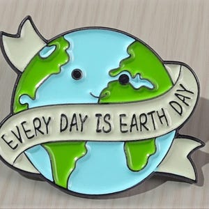 Könnte beinhalten: Ein runder Emaille-Pin mit einer lächelnden Erde mit blauen Ozeanen und grünen Kontinenten. Ein weißes Banner mit dem Text "EVERY DAY IS EARTH DAY" umschließt den Planeten. Der Pin hat eine schwarze Umrandung und ein hellgrünes Band.