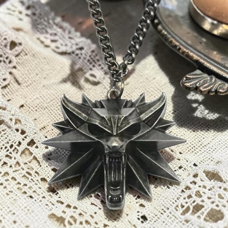 Puede incluir: Collar con colgante plateado con un emblema de cabeza de lobo. El colgante tiene un dise&ntilde;o detallado de una cabeza de lobo con la boca abierta, sujeto a una cadena. El collar es probablemente de metal.