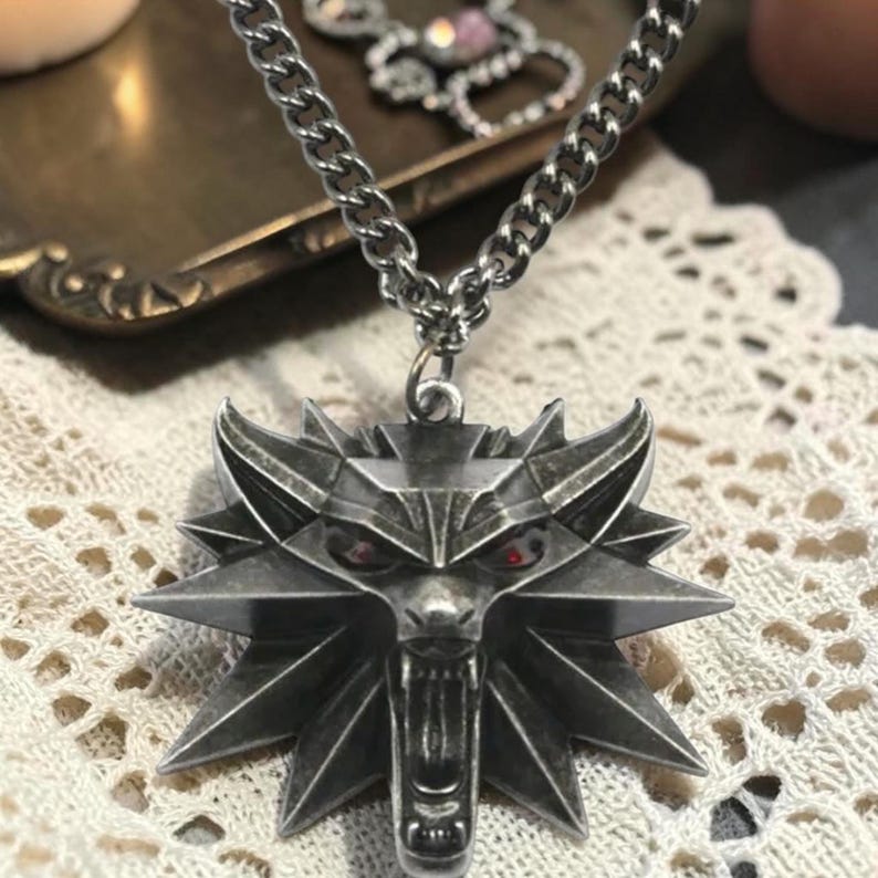 Puede incluir: Un collar con colgante plateado con un dise&ntilde;o de cabeza de lobo con ojos rojos. El colgante est&aacute; unido a una cadena gruesa. El collar se exhibe sobre una superficie de encaje.