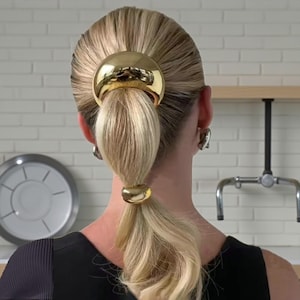 Könnte beinhalten: Blondes Haar, zu einem tiefen Pferdeschwanz gebunden, mit goldfarbenen Haaraccessoires. Das Haar ist glatt und glänzend. Die Frau trägt ein schwarzes Oberteil.