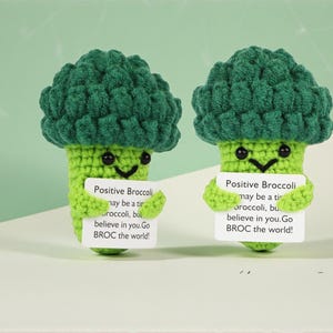 Könnte beinhalten: Zwei handgefertigte gehäkelte Brokkoli-Figuren mit lächelnden Gesichtern, die Schilder mit der Aufschrift "Positive Broccoli" halten. Die Brokkoli-Köpfe sind dunkelgrün und die Stiele hellgrün.
