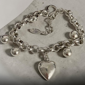 Könnte beinhalten: Ein silbernes Armband mit einem Herzanhänger und kugelförmigen Charms. Das Armband besteht aus verbundenen, abgerundeten Silberstücken. Der Herzanhänger ist glatt und poliert. Das Armband liegt auf einer grauen Oberfläche.