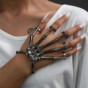 May include: A silver-toned skeleton hand bracelet with rings on each finger. The bracelet is detailed with a skeletal design, including the wrist bones and finger joints. The jewelry is worn on a hand with long, polished fingernails.