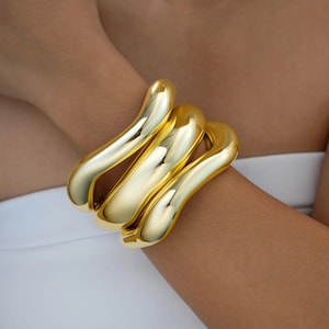 Könnte beinhalten: Ein goldfarbenes Armband mit einem glatten, geschwungenen Design. Das Armband hat eine glänzende Oberfläche und scheint aus Metall zu sein. Das Design zeigt drei verbundene, längliche Schlaufen, die sich um das Handgelenk wickeln. Der Schmuck ist ein Statement-Stück.