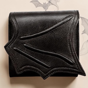 Könnte beinhalten: Schwarze Ledergeldbörse mit einem Fledermausflügel-Design auf der Vorderseite. Die Geldbörse hat eine strukturierte Oberfläche und einen Klappenverschluss. Das Fledermausflügel-Design ist geprägt und hat drei gebogene Linien. Die Geldbörse ist quadratisch.