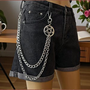 Könnte beinhalten: Silberne Ketten-Accessoire mit einem Pentagramm-Anhänger an schwarzen Jeansshorts. Die Kette besteht aus mehreren Gliedern und hängt am Bein herunter. Die Shorts haben umgeschlagene Manschetten.