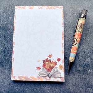 May include: A notepad with a fall-themed design featuring an open book and autumn leaves. The notepad has a decorative border. A patterned pen with black ink rests beside the notepad.